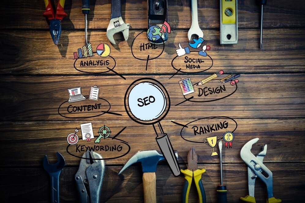 Digital marketing strategy tools illustrated with icons and keywords like SEO, Content, Keywording, Ranking, Design, Social Media, HTML, and Analysis surrounded by various work tools on a wooden background.